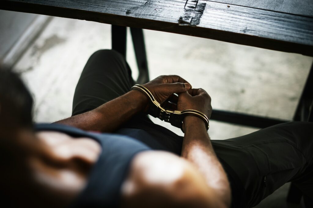A person sits with their hands cuffed together in front of them beneath a rough wooden table, viewed from above, conveying detention or arrest in a stark, dimly lit setting.
