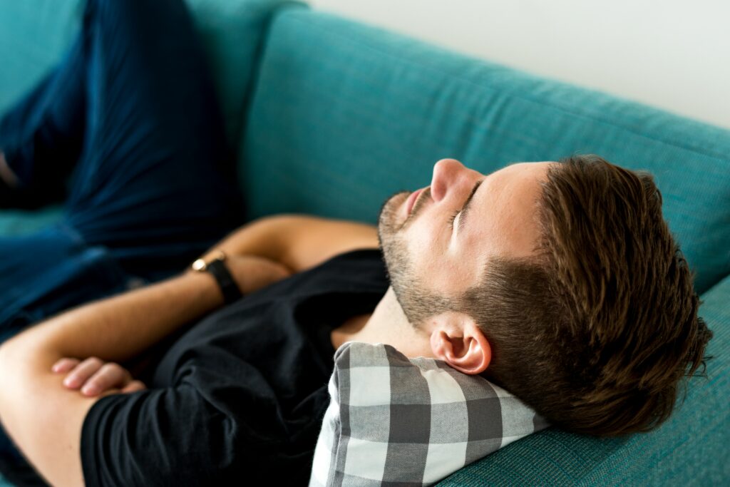 Adult man lying on a teal couch with eyes closed, arms folded across his torso, head resting on a checkered pillow, appearing to rest or sleep in a calm indoor setting.