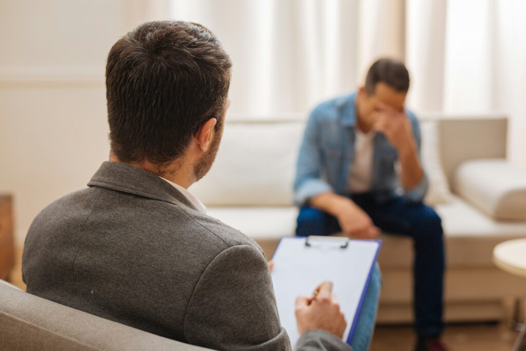 Man seated on a couch with head lowered and hand covering his face during a counseling session, while a therapist in the foreground takes notes on a clipboard in a softly lit room.