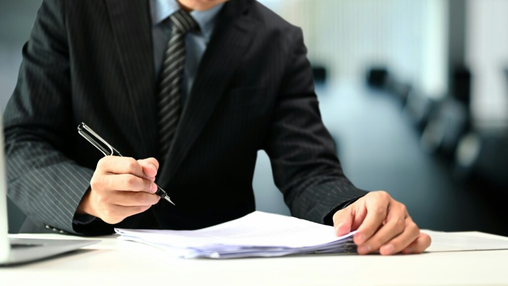Business professional in a suit reviews and signs a stack of documents at a desk, holding a pen during a formal paperwork process.