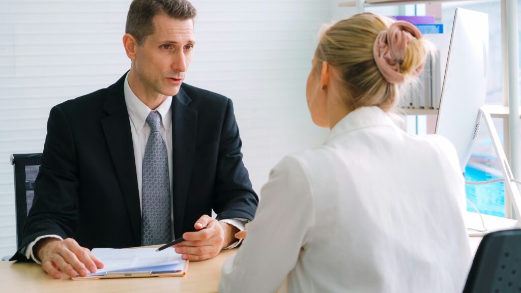 A professionally dressed man in a suit sits at a desk holding a pen and paperwork while speaking with a woman seated across from him in an office, suggesting a formal consultation or legal meeting.
