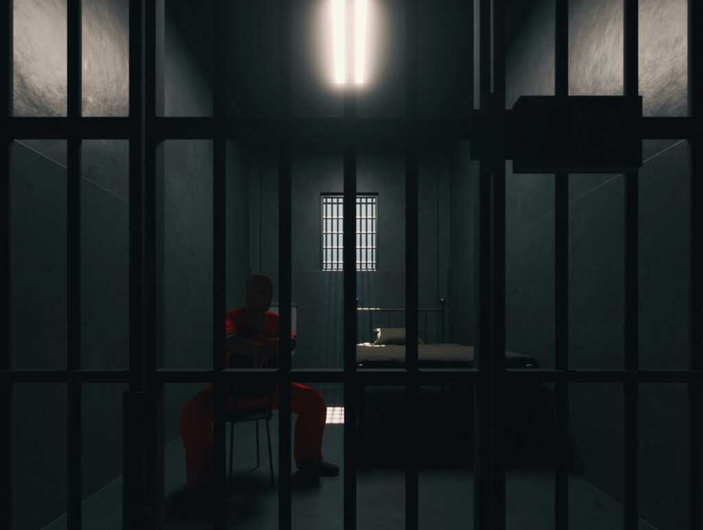 A dimly lit prison cell viewed through thick vertical metal bars, with a solitary figure in an orange jumpsuit sitting hunched on a chair near the center of the room, head lowered and arms resting on their knees. A narrow barred window at the back lets in a faint beam of light that falls across a simple metal bed with a thin mattress and pillow, emphasizing the stark, confined space, cold concrete walls, and heavy shadows that create a somber, isolating atmosphere.