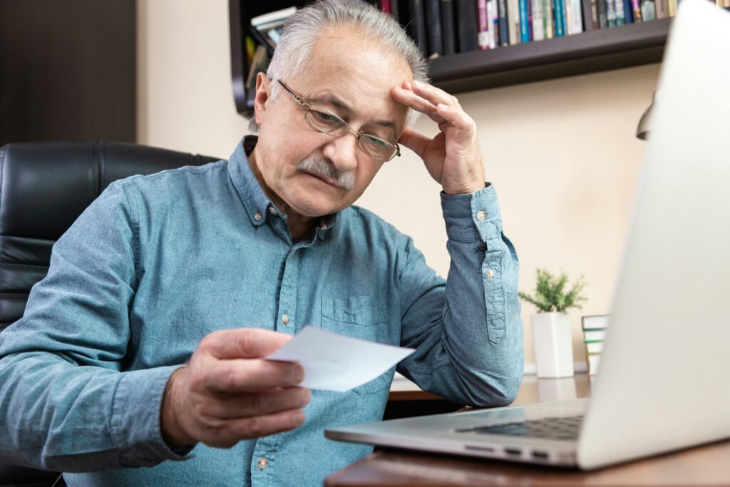 Older man sitting at a desk reviews a small document with a concerned expression while working on a laptop in a home office with bookshelves in the background.